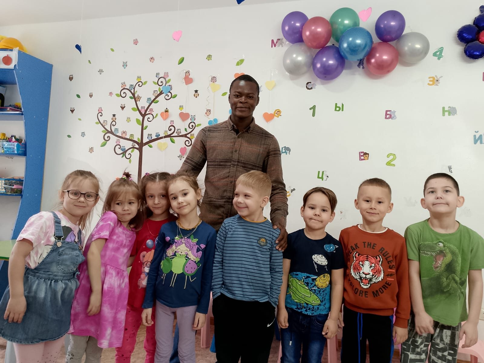 Emmanuel with a full class of young learners in a bright room decorated with balloons and letters